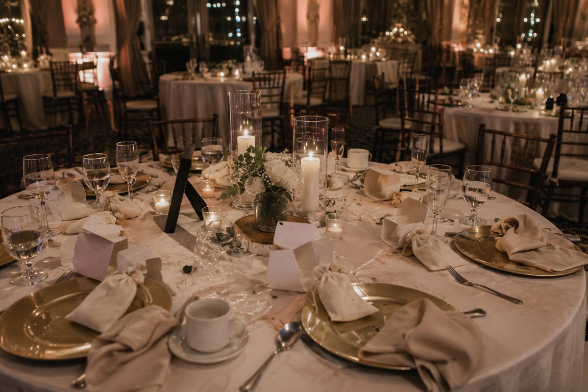 A candlelit wedding reception ballroom late at night, gold chargers still set with linen napkins and drawstring favor pouches left unclaimed at every place setting